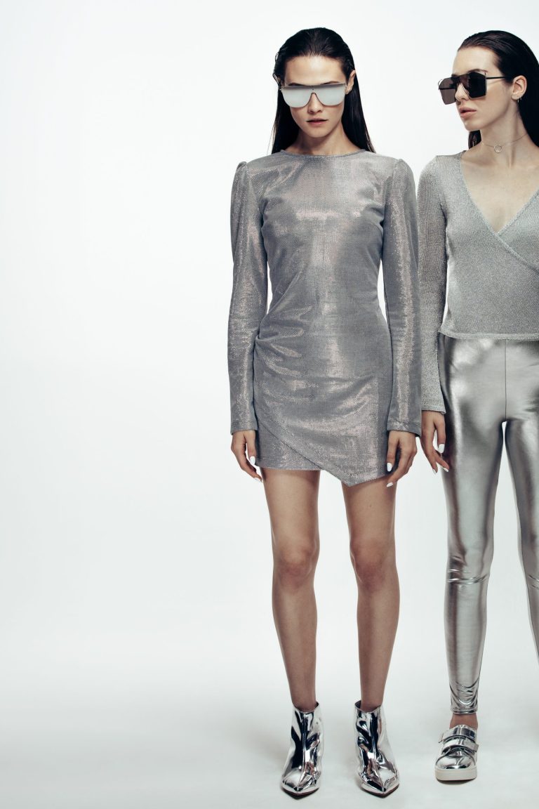 Full length portrait of two young women wearing silver outfit and sunglasses standing over grey background. Female model in futuristic look in studio.